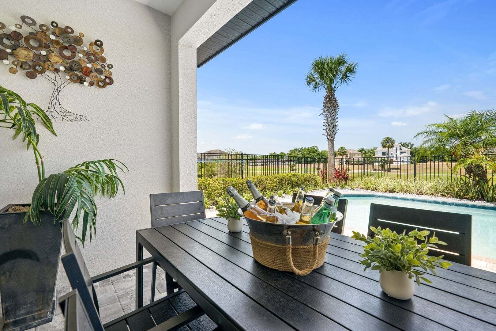 A patio with a table, chairs, a decorative wall, and a view of a swimming pool and palm trees.