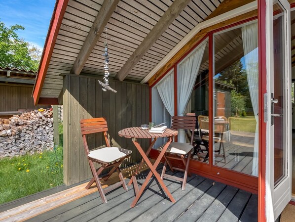 Um deck de madeira com uma mesa e cadeiras, um sino de vento pendurado e uma porta de vidro que dá para uma área externa.