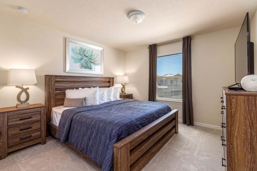 Une chambre à coucher avec un lit en bois, deux tables de chevet avec des lampes, une grande fenêtre avec des rideaux et un luminaire au plafond.