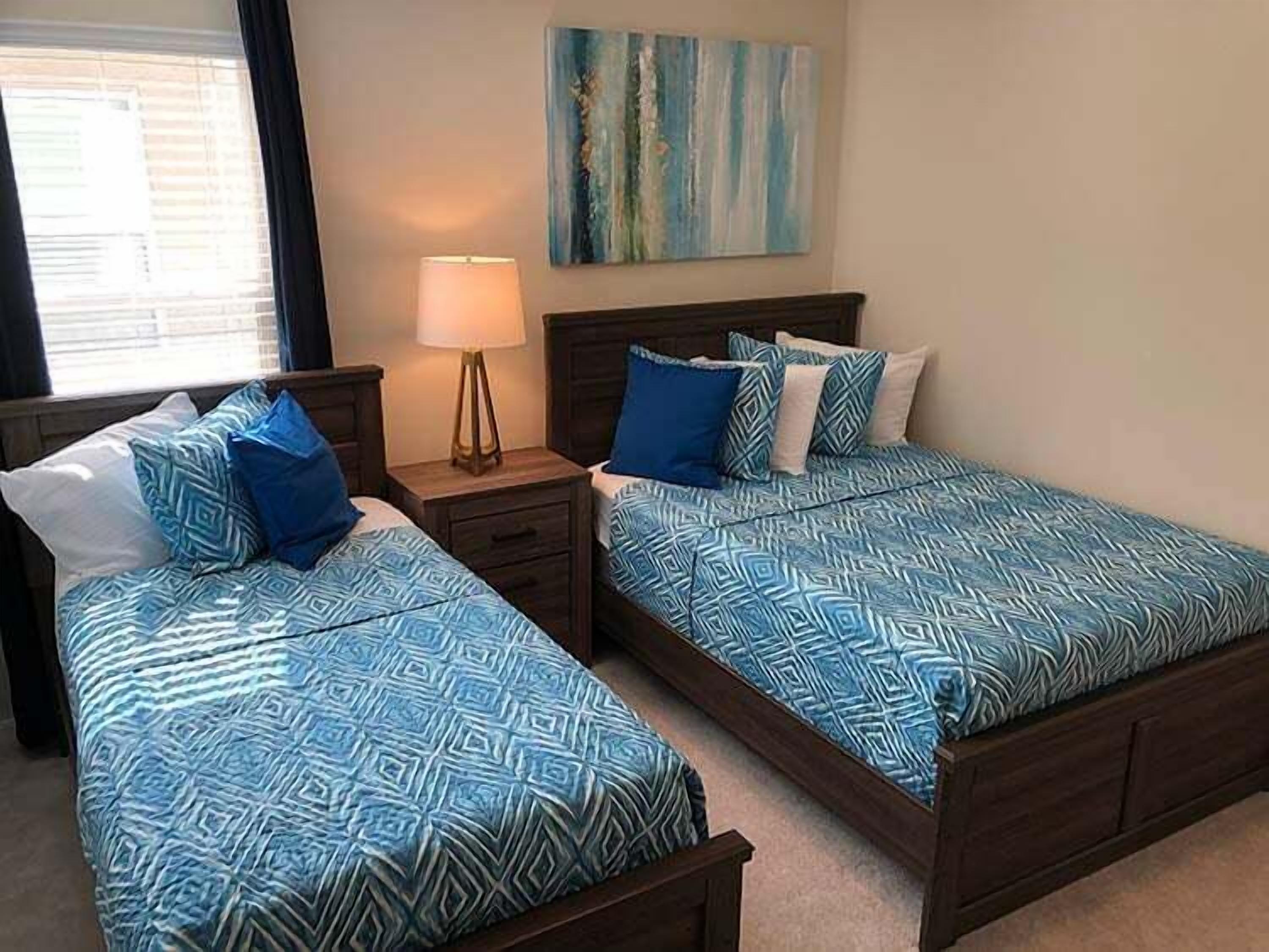 A hotel room with two beds, a bedside lamp, a painting on the wall, and a window with blinds.