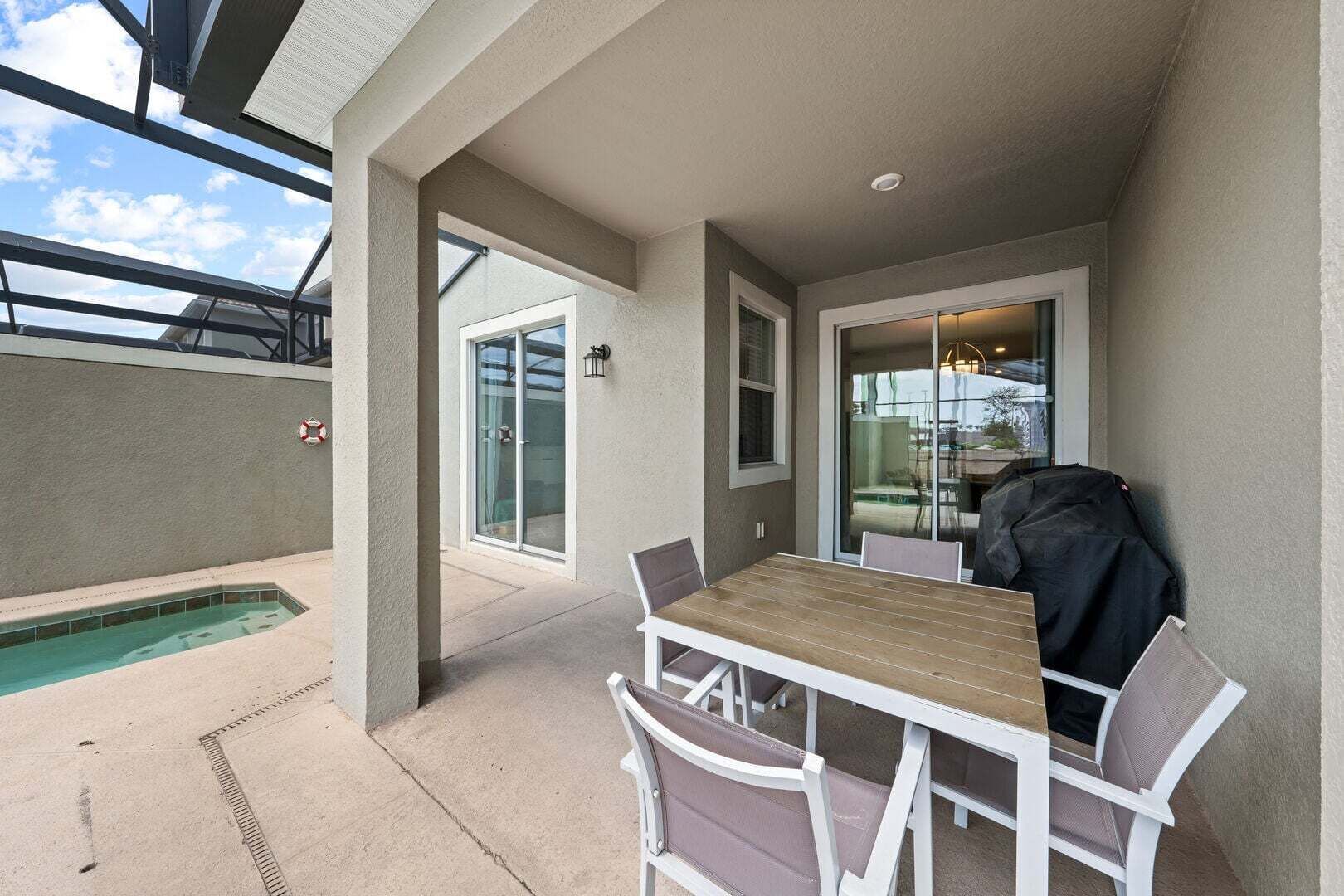 A covered patio with a pool, a table with chairs, and a grill.