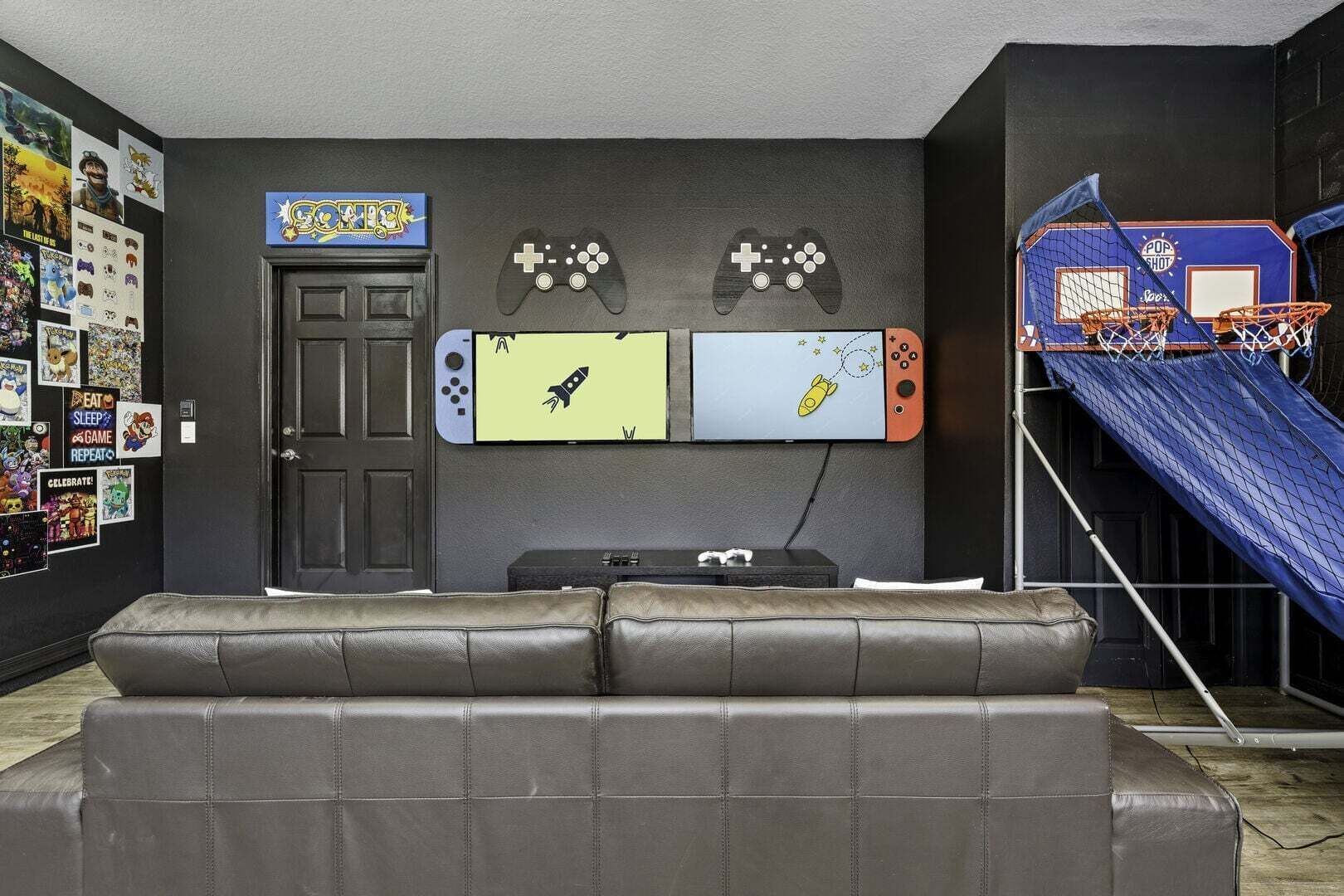 Une pièce comprenant un canapé, une porte, des éléments de décoration murale, notamment des manettes de jeu, et un panier de basket.
