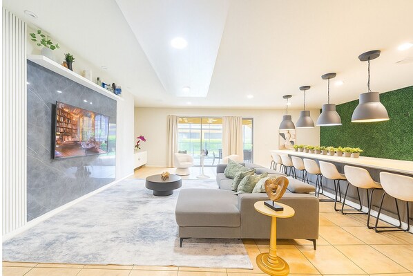 A modern living room with a sofa, coffee table, and a TV. There is a dining area with chairs and pendant lights. - Comfortable Vacation Home - Great Resort Pool/spa Sleeps 20 Ww101 (Kissimmee)
