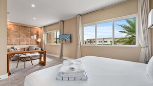 Ein Hotelzimmer mit Bett, Schreibtisch, Stuhl, Fernseher und einem Fenster mit Blick auf Palmen.