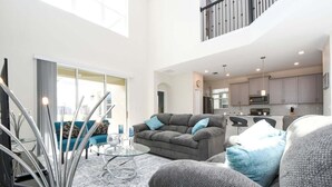 A spacious living area with a grey sofa, a glass coffee table, and a blue armchair. There is a kitchen in the background with modern appliances. - Champions Gate Gem Pool, Spa, Near Parks (Davenport)