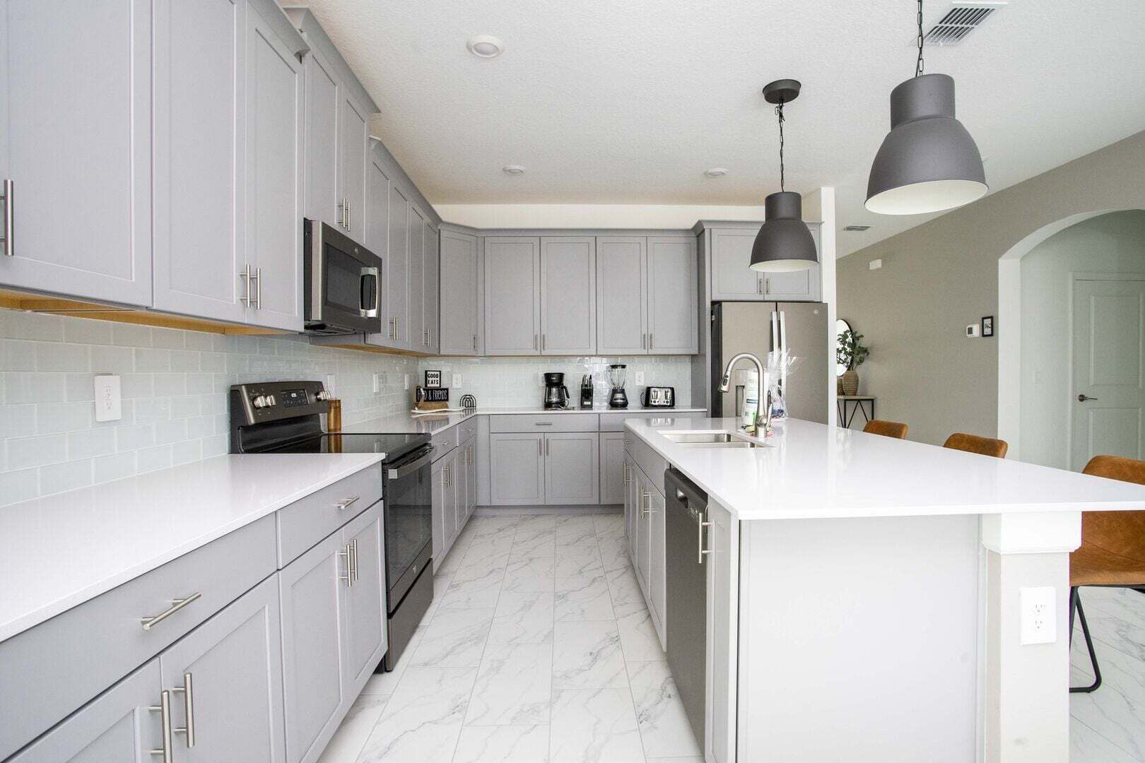 A modern kitchen with grey cabinets, a white countertop, a built-in oven, a microwave, a coffee maker, and a large island with a sink.