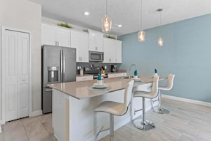 A modern kitchen with white cabinets, stainless steel appliances, a central island with bar stools, and pendant lighting. - Lovely Family Home - 8 Min to Disney! (Kissimmee)