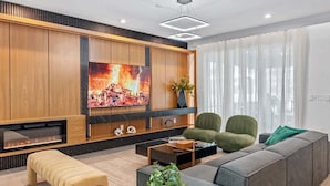 Ein modernes Wohnzimmer mit großem Flachbildfernseher, einer Couch, zwei Sesseln, einem Couchtisch und einem Kamin.