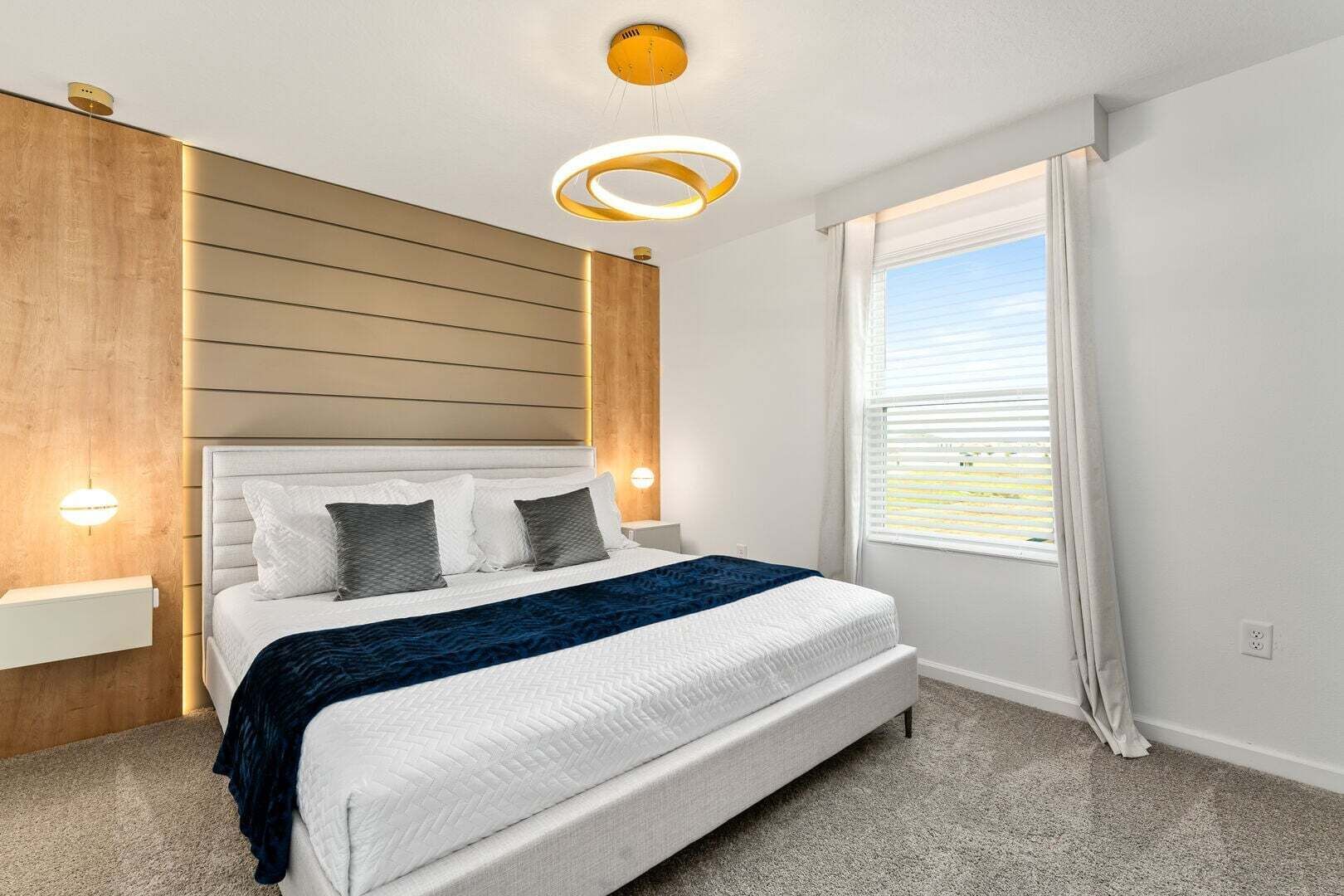 A modern bedroom with a large bed, built-in wooden panel wall, and a window with blinds.