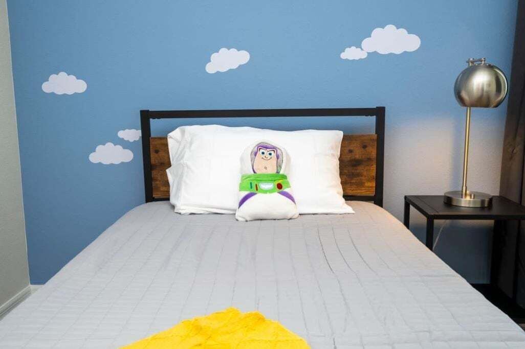 A bedroom with a bed, a pillow with a character, a bedside lamp, and a wall with cloud decals.