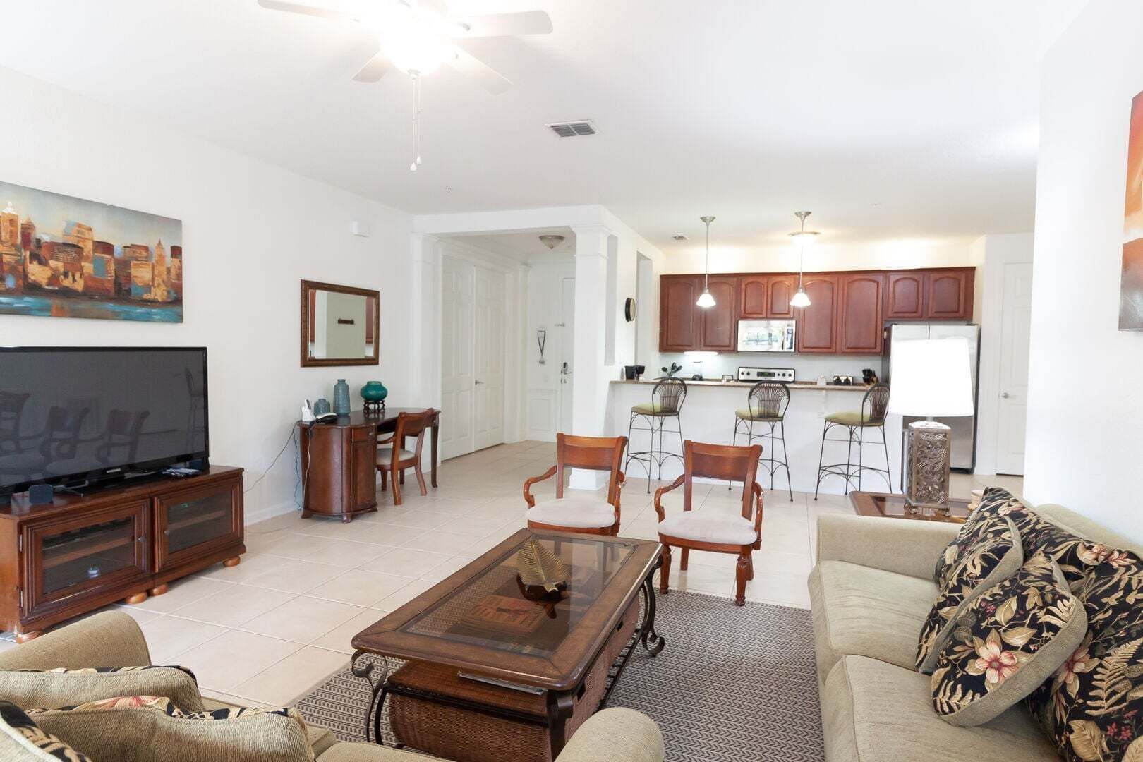 Una sala de estar con televisor, muebles de madera, comedor y cocina con alacenas y taburetes de bar.