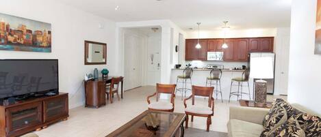 Una sala de estar con televisor, muebles de madera, comedor y cocina con alacenas y taburetes de bar.