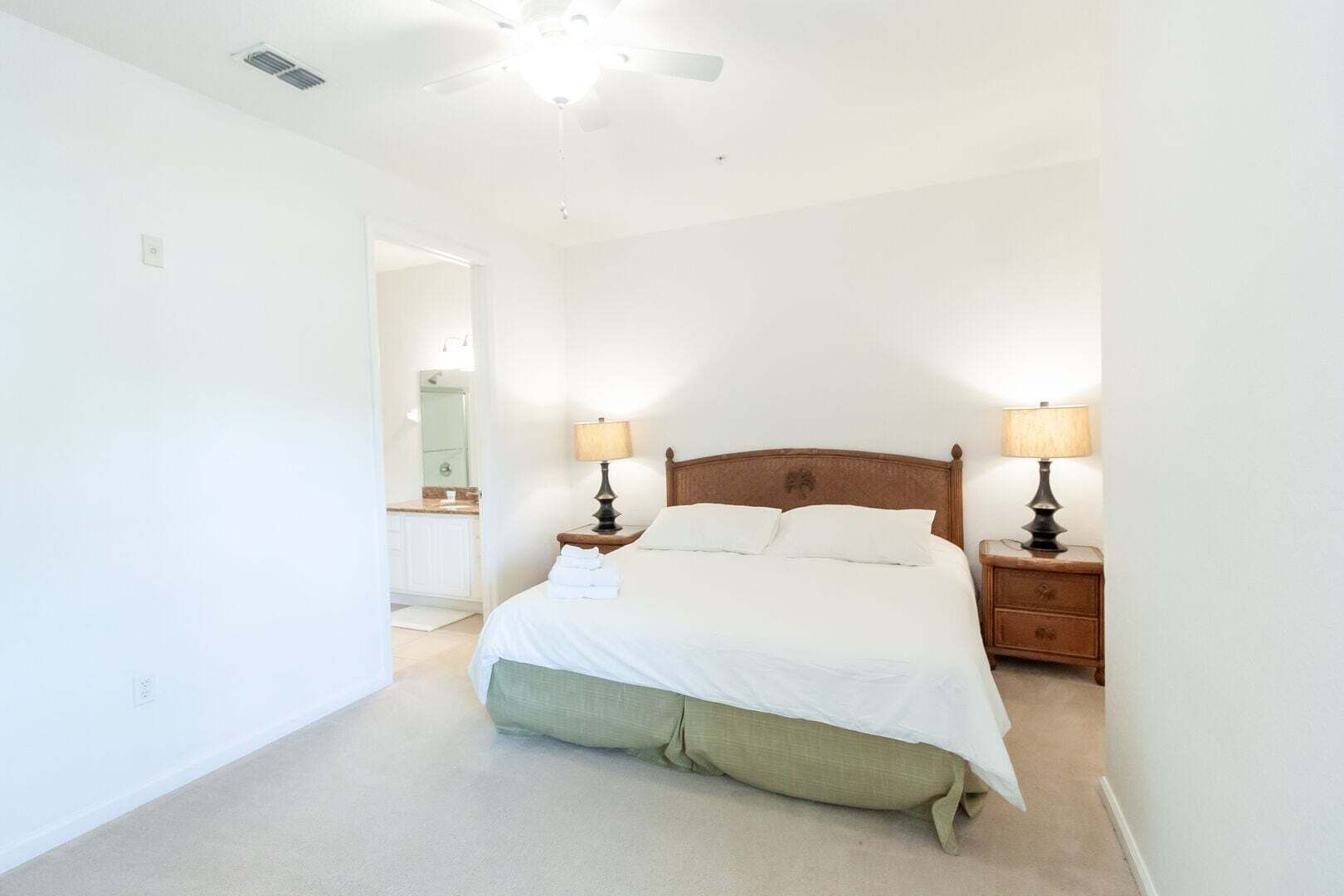 Une chambre à coucher avec un lit, deux tables de chevet avec des lampes, un ventilateur de plafond et une salle de bain visible à travers une porte ouverte.