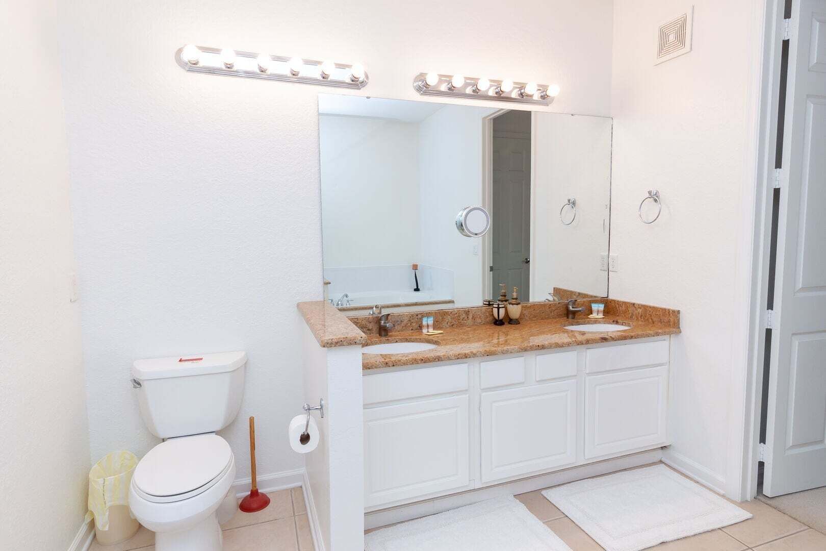 A bathroom with a white toilet, two sinks with granite countertops, a large mirror with lights, and white cabinets.