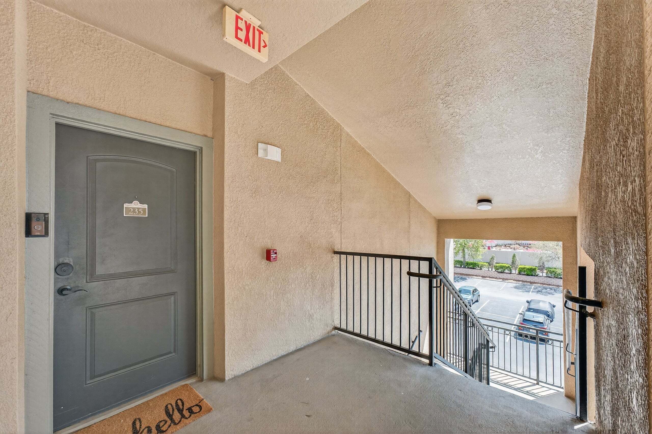 Entrada de un edificio con una puerta gris marcada '235', un letrero de salida, un detector de incendios y vistas a un estacionamiento con coches.