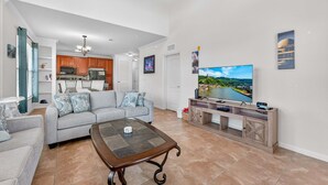 Una sala de estar con un sofá gris, una mesa de centro, un televisor y una cocina al fondo.