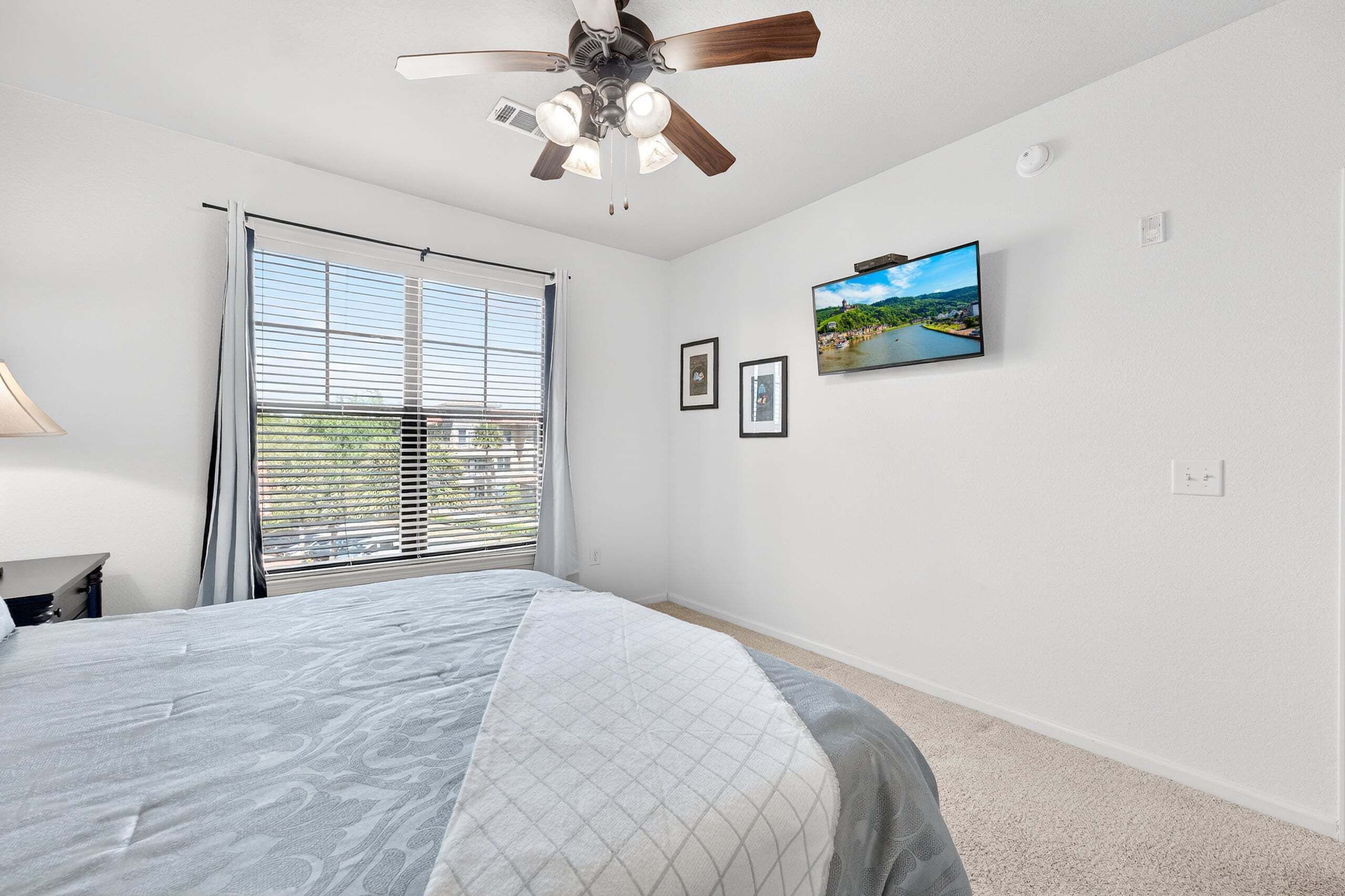 A bedroom with a bed, ceiling fan, window with blinds, wall-mounted TV, and framed pictures.