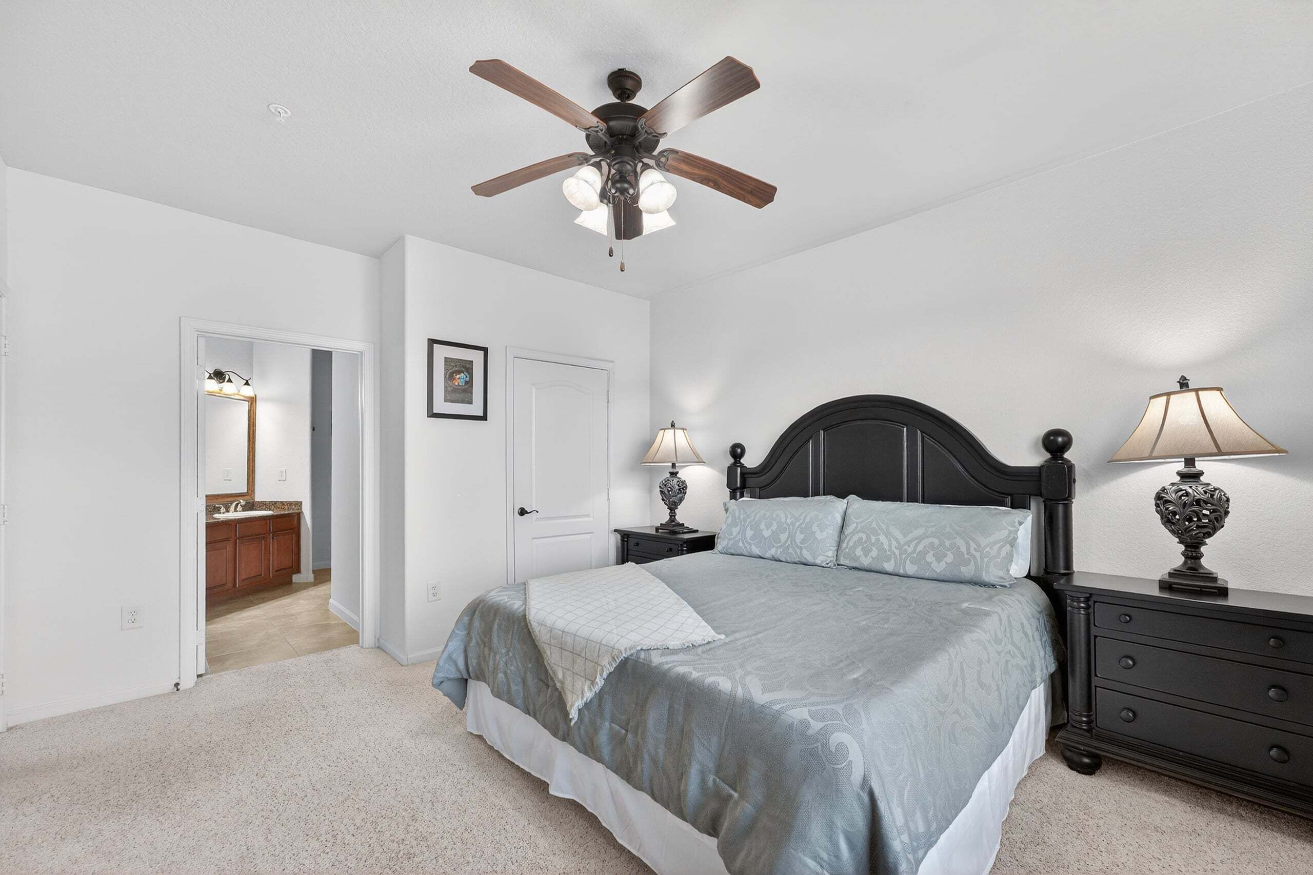 A bedroom with a bed, a ceiling fan, bedside lamps, a nightstand, and a door leading to a bathroom.