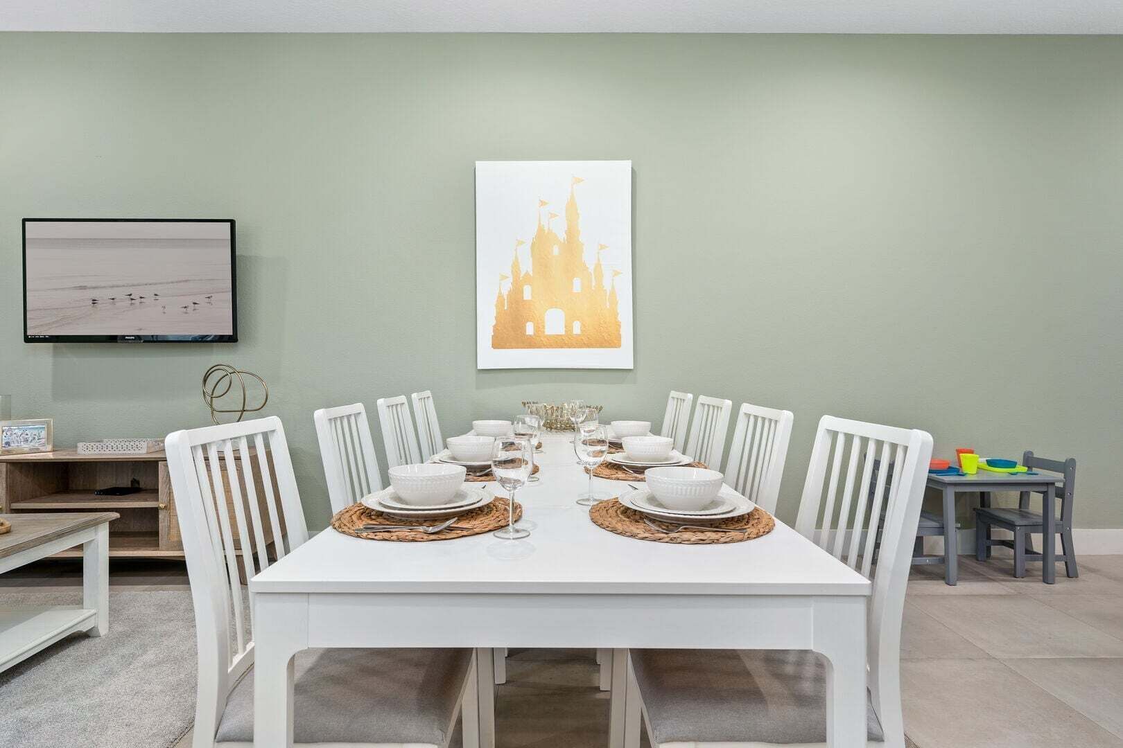 Uma sala de jantar com uma mesa branca posta para oito pessoas, rodeada por cadeiras brancas. Uma moldura com um quadro está pendurada na parede acima da mesa.