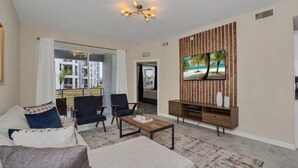 A living room with a sofa, armchairs, a coffee table, a TV on a wooden wall, and a view of a building outside. - Precious Condo 8min to Disneyworld Resort Access St105 (Kissimmee)
