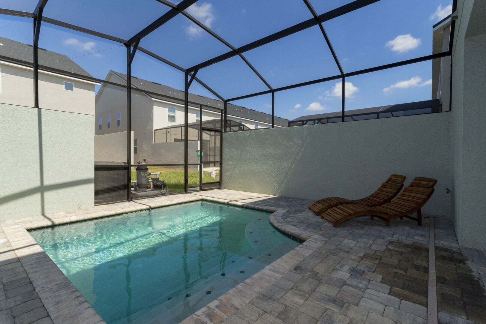Une piscine entourée de parois en verre, deux chaises longues en bois et une terrasse en briques.