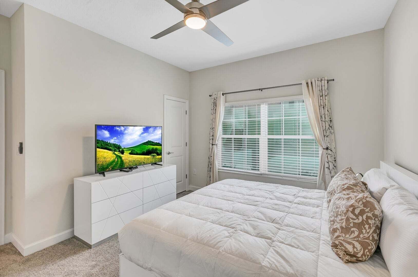 Ein Schlafzimmer mit einem Bett, einem Fernseher auf einer weißen Kommode, einem Deckenventilator und einem Fenster mit Vorhängen.