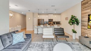 A modern living room with a grey sofa, white chairs, a dining area, and a kitchen with white cabinets and pendant lights. - Private Pool! 8 Min to Disney! (Kissimmee)