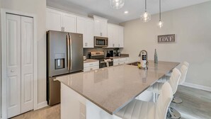 A modern kitchen with white cabinetry, stainless steel appliances, a large island with seating, pendant lighting, and a wall-mounted microwave. - Private Pool! 8 Min to Disney! (Kissimmee)