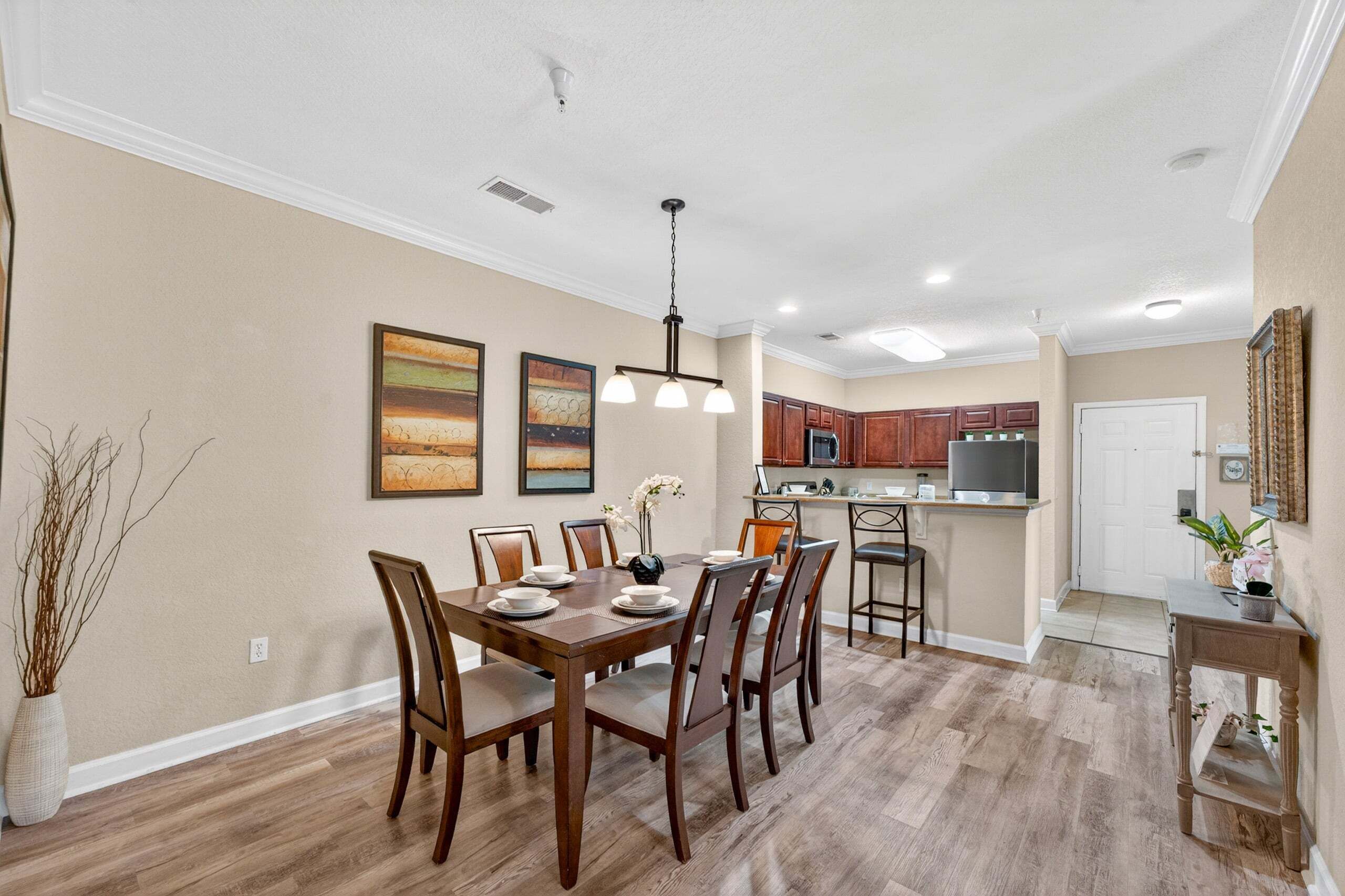 A dining area with a wooden table and chairs, a kitchen in the background, and a white door.