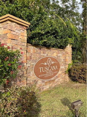 A stone wall with a circular sign that reads 'TUSCANA ORLANDO'S MEDITERRANEAN INSPIRED GARDENS CONDOMINIUMS.' - Condo 2BR With Lanai & Shared Pool, hot Tub, gym (Davenport)