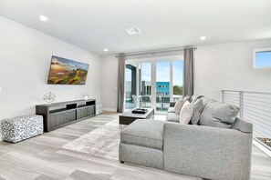 Ein modernes Wohnzimmer mit einem grauen Sofa, einem gemusterten Hocker, einem Holztisch und einem an der Wand befestigten Fernseher.