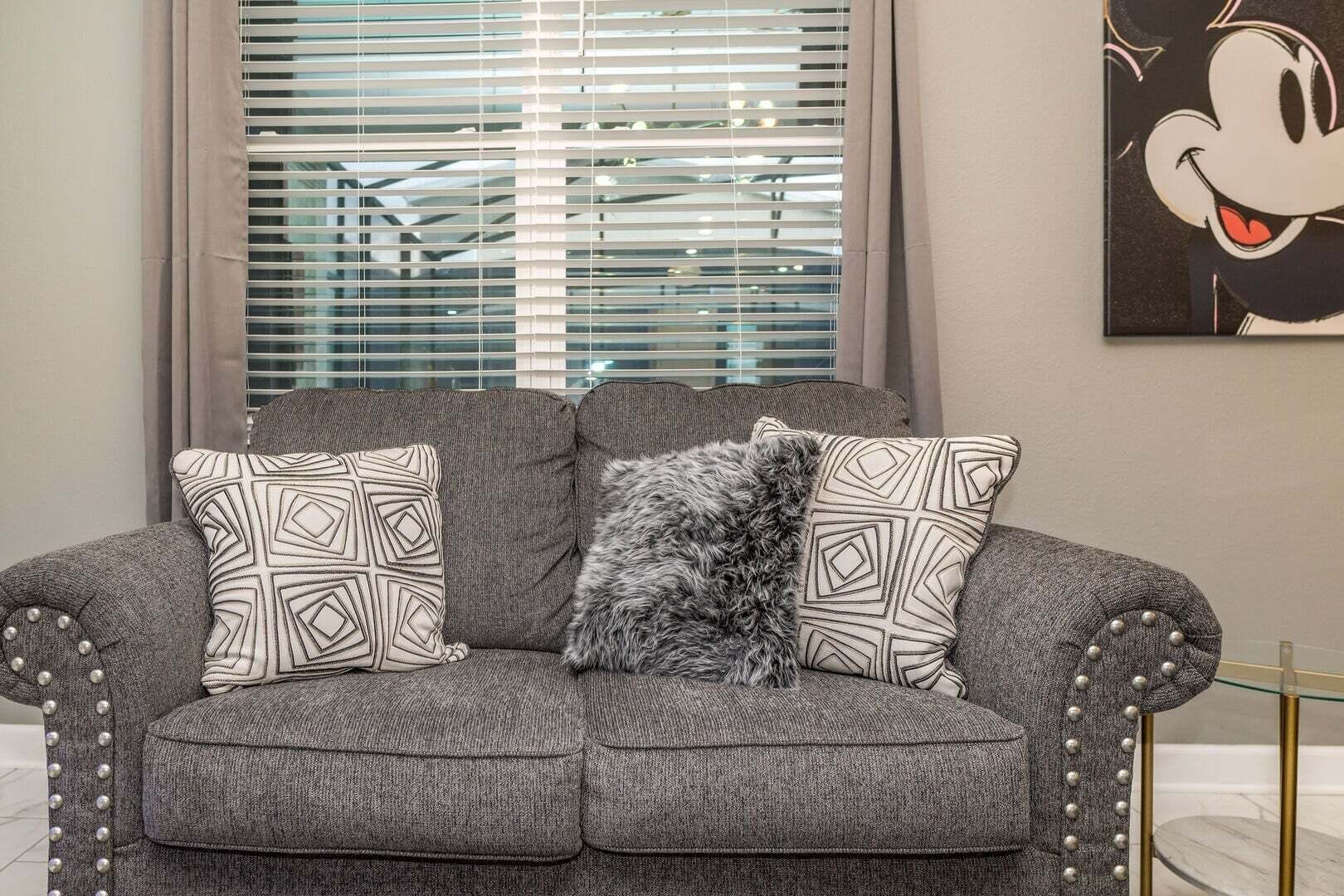 En grå polstret sofa med mønstrede puder, et blødt tæppe og et billede af Mickey Mouse på væggen.