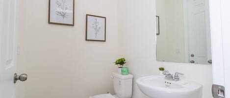 Une salle de bain comprenant des toilettes, un lavabo, deux illustrations botaniques encadrées accrochées au mur, une petite plante posée sur le réservoir des toilettes et un miroir au-dessus du lavabo.