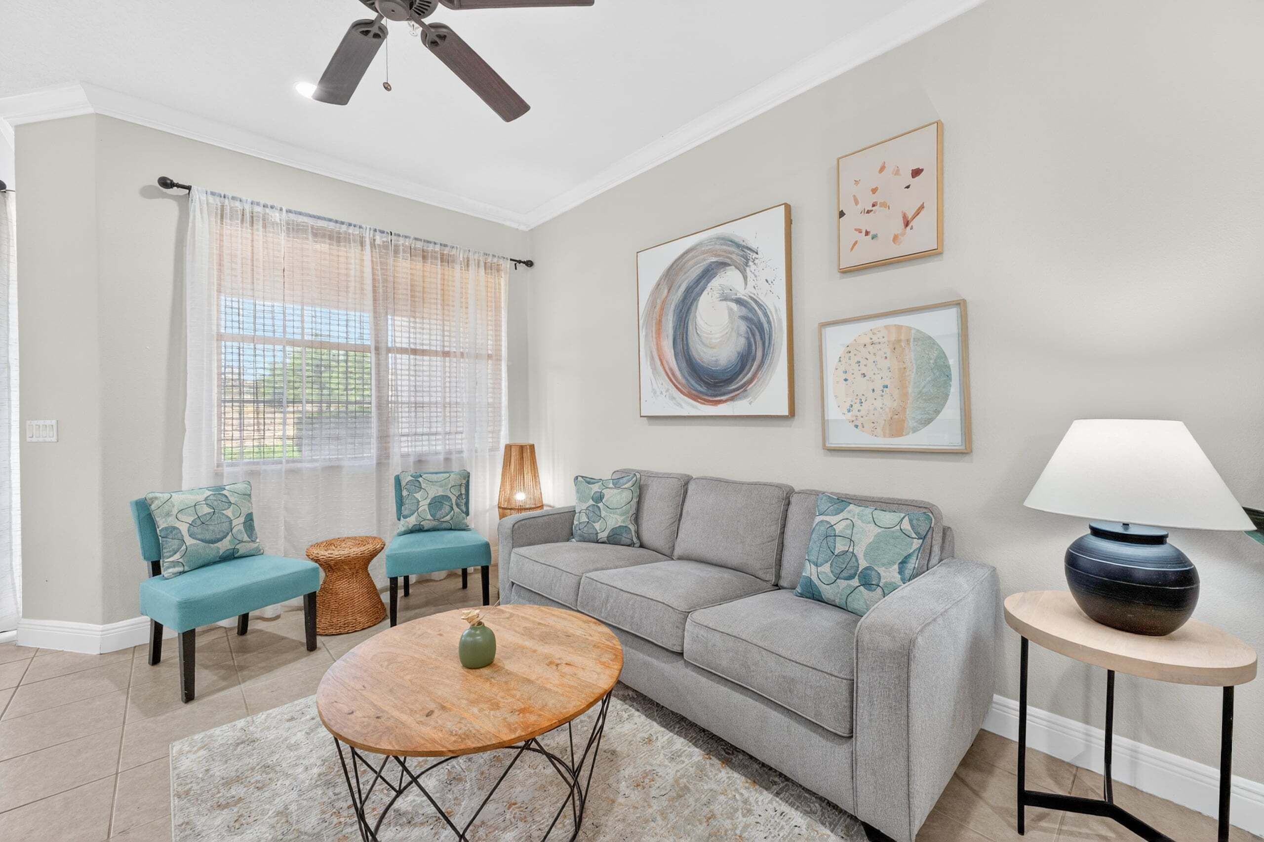A living room with a grey sofa, teal armchairs, a round coffee table, a ceiling fan, and artwork on the wall.