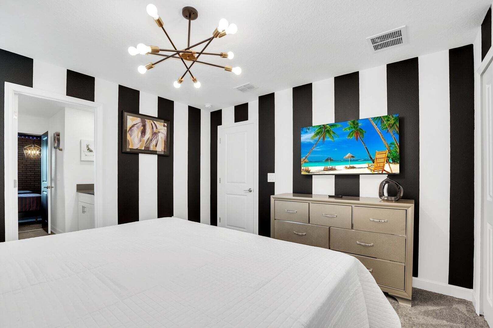 Ein Schlafzimmer mit einem großen Bett, einem Fernseher auf einer Kommode, einer Deckenleuchte und einer Tür zu einem anderen Raum.