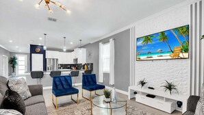 A modern living room with a large TV, two blue armchairs, a glass coffee table, and a kitchen in the background. - Magical Mickey Home 8min->disney! (Kissimmee)