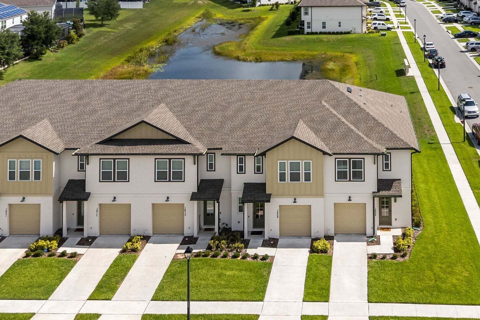 A row of two-story houses with attached garages, a paved driveway, and a small water feature in the background.