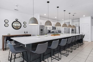 A modern kitchen with a central island, bar stools, pendant lights, and a dining area with chairs and a table. - Unique Masterpiece 15 En-suites! (Kissimmee)