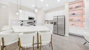 Een moderne keuken met witte kasten, roestvrijstalen apparatuur, een centraal eiland met barkrukken, hanglampen en een decoratieve plank met keramieken objecten.