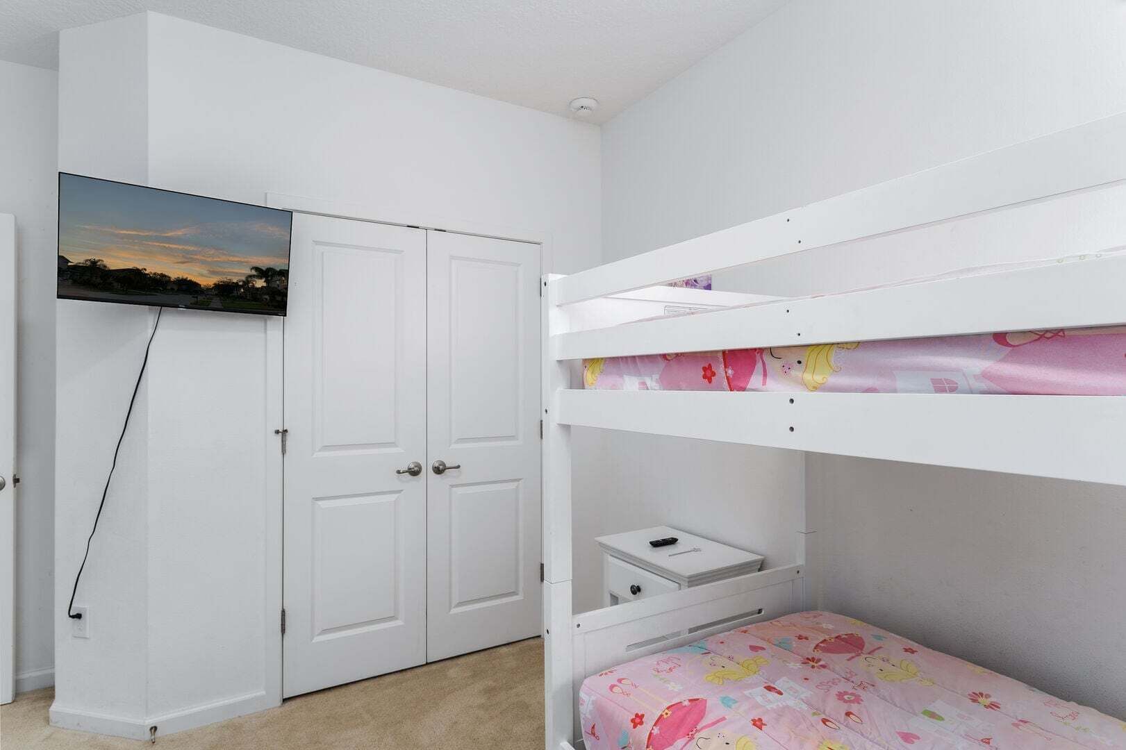 Un lit superposé avec une couvre-lit à motifs floraux roses, une armoire blanche surmontée d’un téléviseur, et une porte avec un volet fermé.