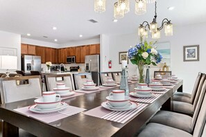 A dining area with a long table set for a meal, kitchen in the background, and a chandelier above. - Lakeview Home Sleeps 25 (Kissimmee)