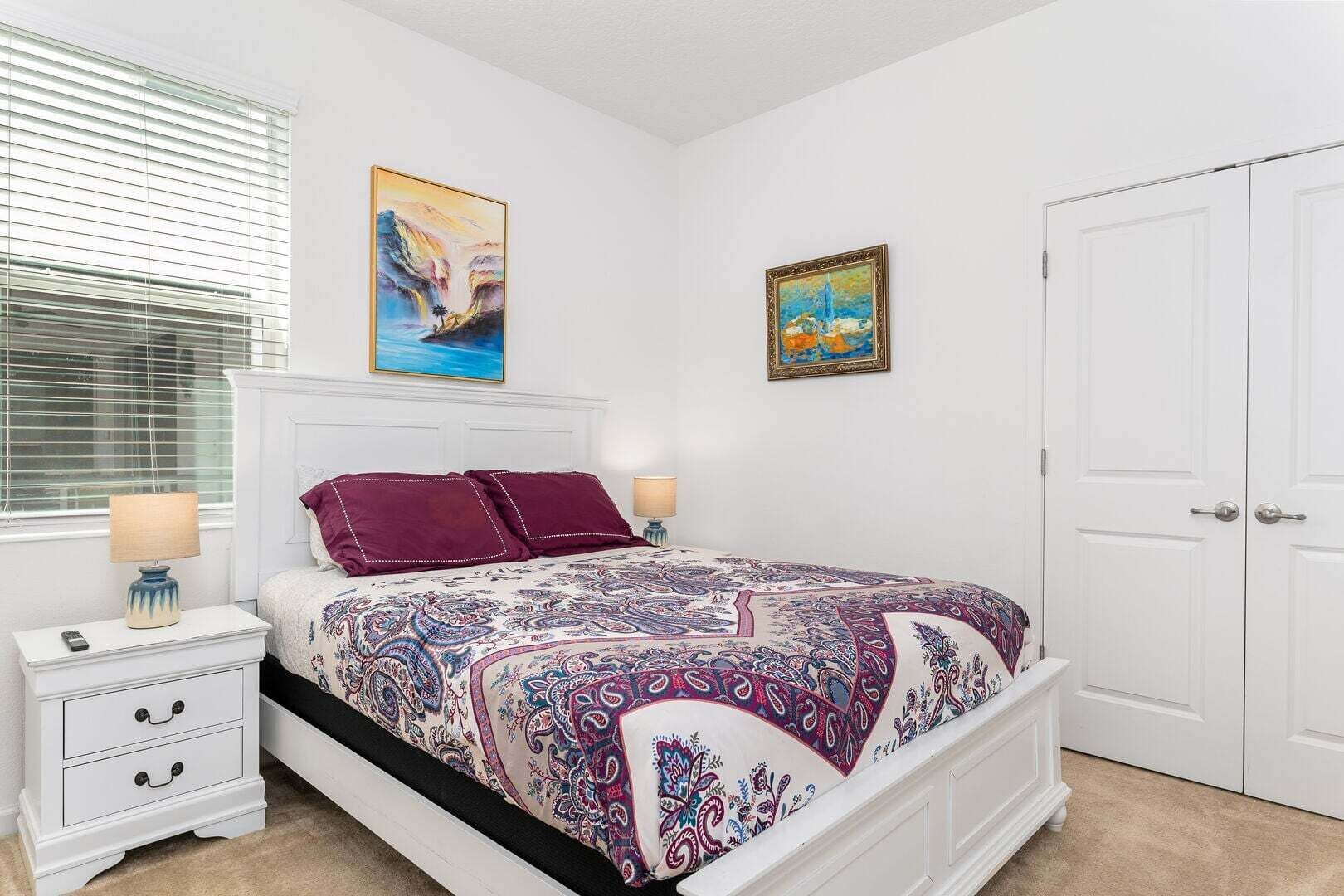 A bedroom with a bed, two bedside tables, a nightstand, a window with blinds, and two doors.