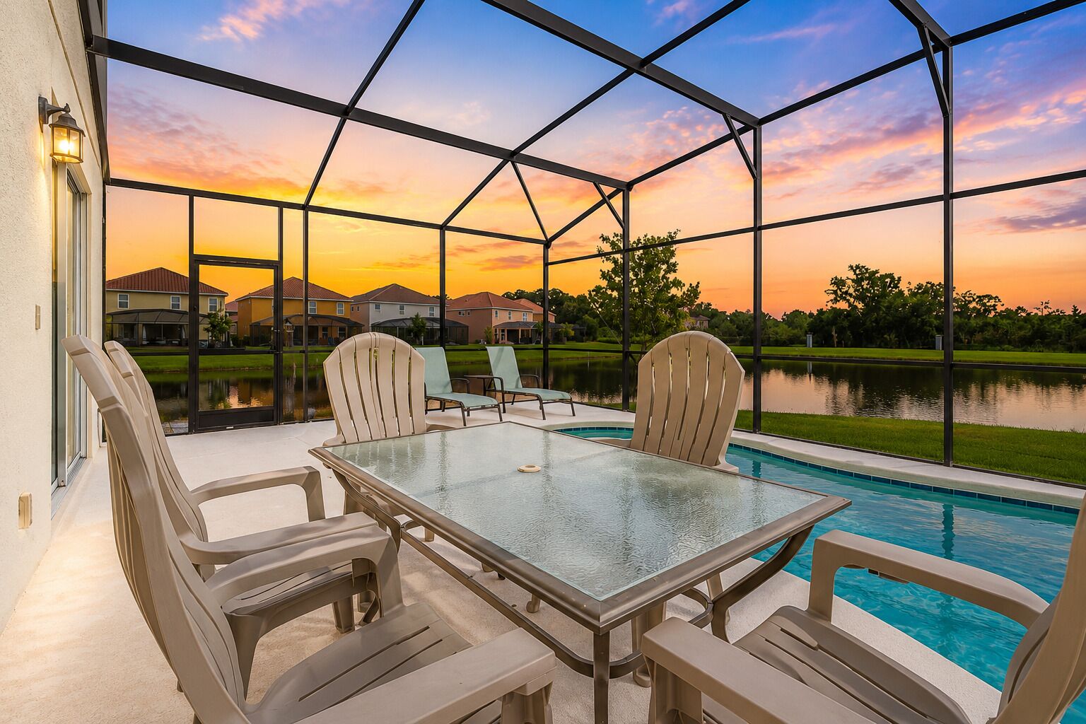 A covered outdoor area with a glass table and chairs, a swimming pool, and a view of houses and a lake at sunset.