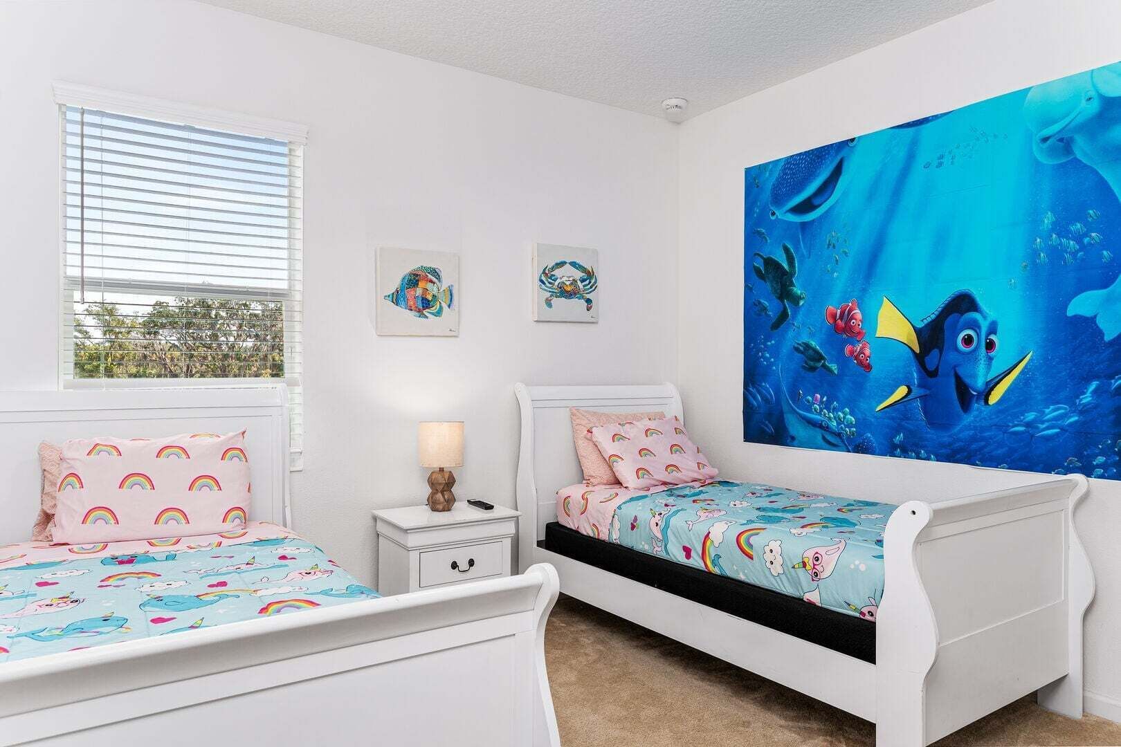A bedroom with two beds, a window with blinds, a nightstand with a lamp, and a large wall mural.