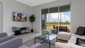 Een moderne woonkamer met een grote televisie, een glazen salontafel en een bank. Er is een balkon met uitzicht op een park en een gebouw.