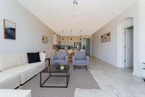 Una sala de estar moderna con un sofá, dos sillones, una mesita de centro y una cocina al fondo.