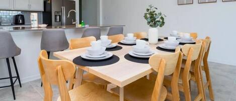 Un comedor moderno con una mesa de madera dispuesta para 10 personas, rodeada de sillas grises. Se ve una cocina con electrodomésticos modernos al fondo.