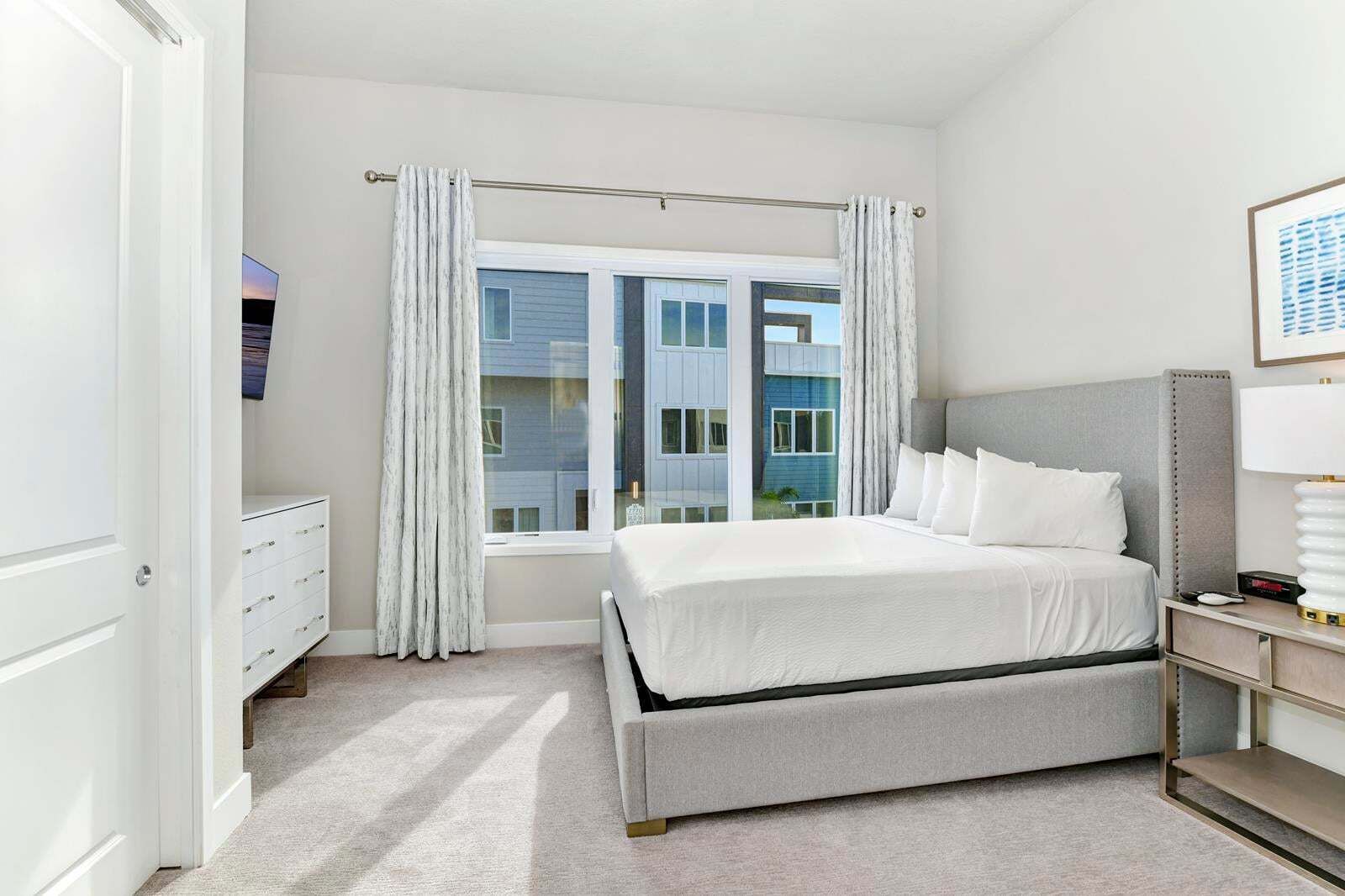 Un dormitorio con cama, mesita de noche, lámpara, ventana con cortinas y vistas a edificios.