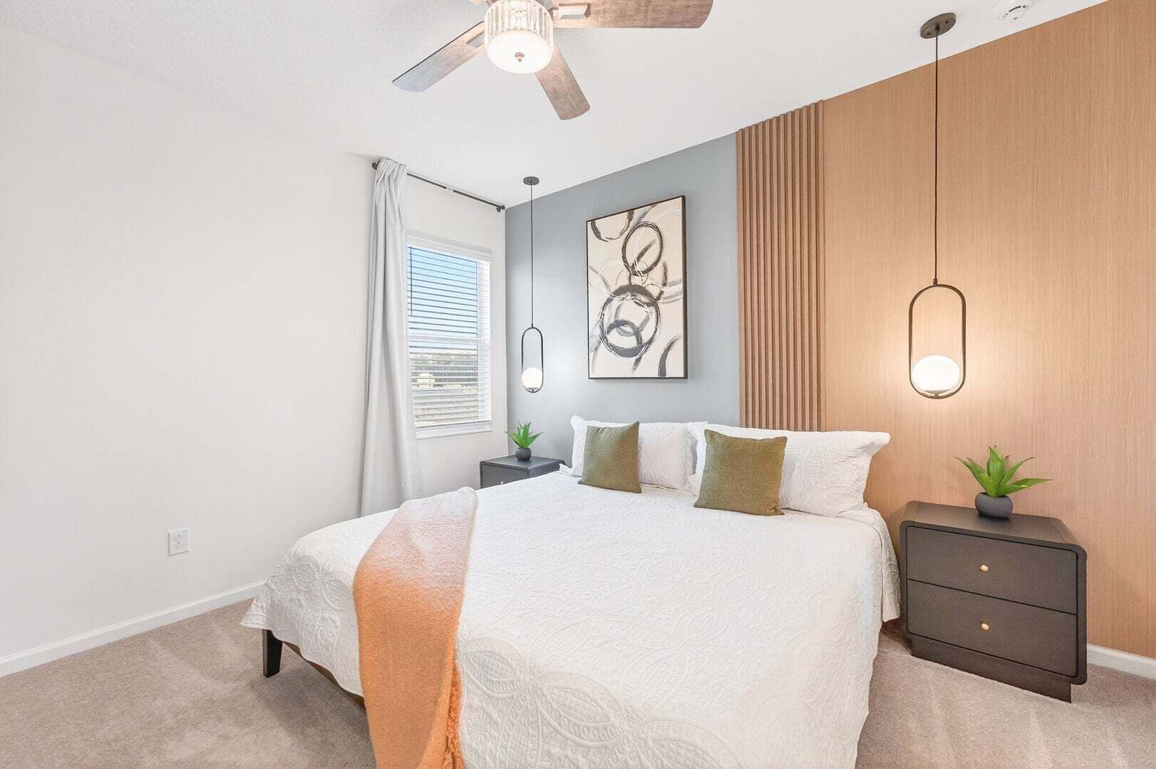 A bedroom with a bed, bedside tables, a ceiling fan, a window with blinds, and a wall-mounted light fixture.