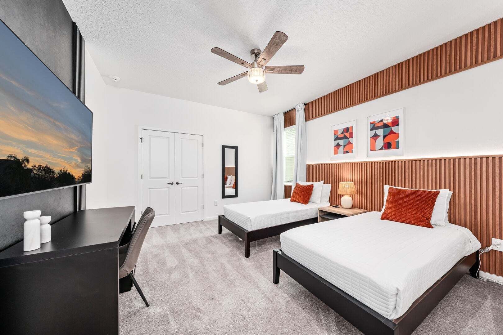 A modern hotel room with a large bed, two smaller beds, a desk, a ceiling fan, and artwork on the walls.